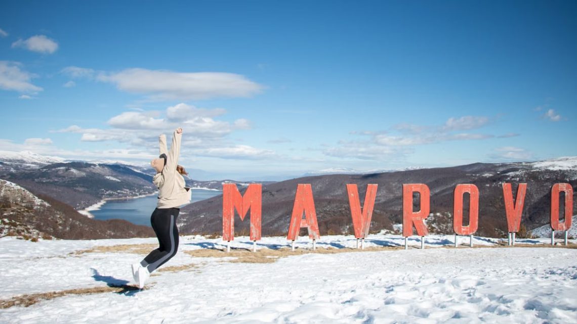 Woman jumping cheerfully in the snow beside Mavrovo sign in North Macedonia.