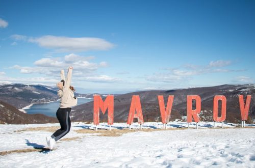 Woman jumping cheerfully in the snow beside Mavrovo sign in North Macedonia.