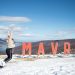 Woman jumping cheerfully in the snow beside Mavrovo sign in North Macedonia.